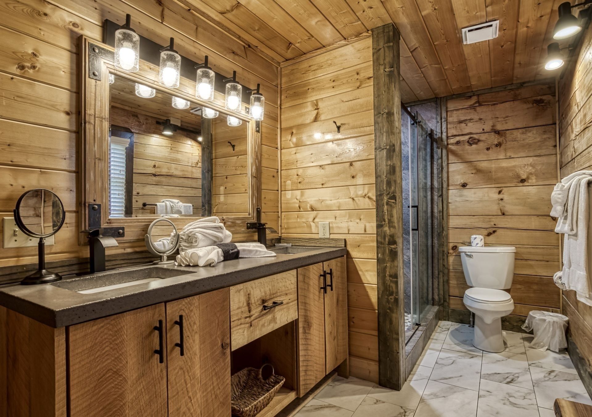 Un baño de cabaña de madera con tocador de madera, lavamanos, espejo con luces, inodoro y zona de ducha.