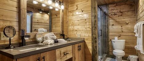 Un baño de cabaña de madera con tocador de madera, lavamanos, espejo con luces, inodoro y zona de ducha.
