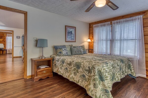 A bedroom with a wooden floor, a bed with floral bedding, a nightstand with a lamp, and a ceiling fan.