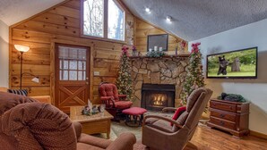 A cozy log cabin interior with a stone fireplace, wooden walls, and a Christmas tree. - Er46 - Cherith Brook - Great Location. Just Across From Swimming Pool! 2 Bedroom Cabin by RedAwning (Pigeon Forge)