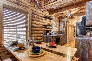 A cozy log cabin interior with a wooden dining table set for four, a kitchen area with a stove and sink, and a window with blinds.