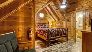 Un dormitorio acogedor en el ático, con techo inclinado de madera, una cama con colcha, un extintor y una pequeña ventana.