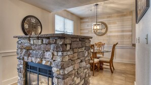 A stone fireplace with a metal screen, a dining area with wooden chairs and a table, and a wall clock. - Owl See You 3205 - Close To Dollywood And Overlooking Gatlinburg Golf Course 2 Bedroom Condo by RedAwning (Pigeon Forge)