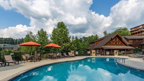 A swimming pool with lounge chairs and umbrellas, surrounded by trees and a wooden building. - Owl See You 3205 - Close To Dollywood And Overlooking Gatlinburg Golf Course 2 Bedroom Condo by RedAwning (Pigeon Forge)