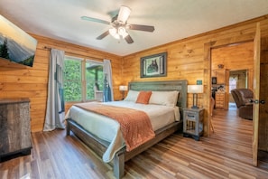 A wooden cabin bedroom with a bed, a ceiling fan, a window with curtains, and a door leading to another room. - Er37 - Hi-bear-nate Great Location..close To Town 1 Bedroom Cabin by RedAwning (Pigeon Forge)