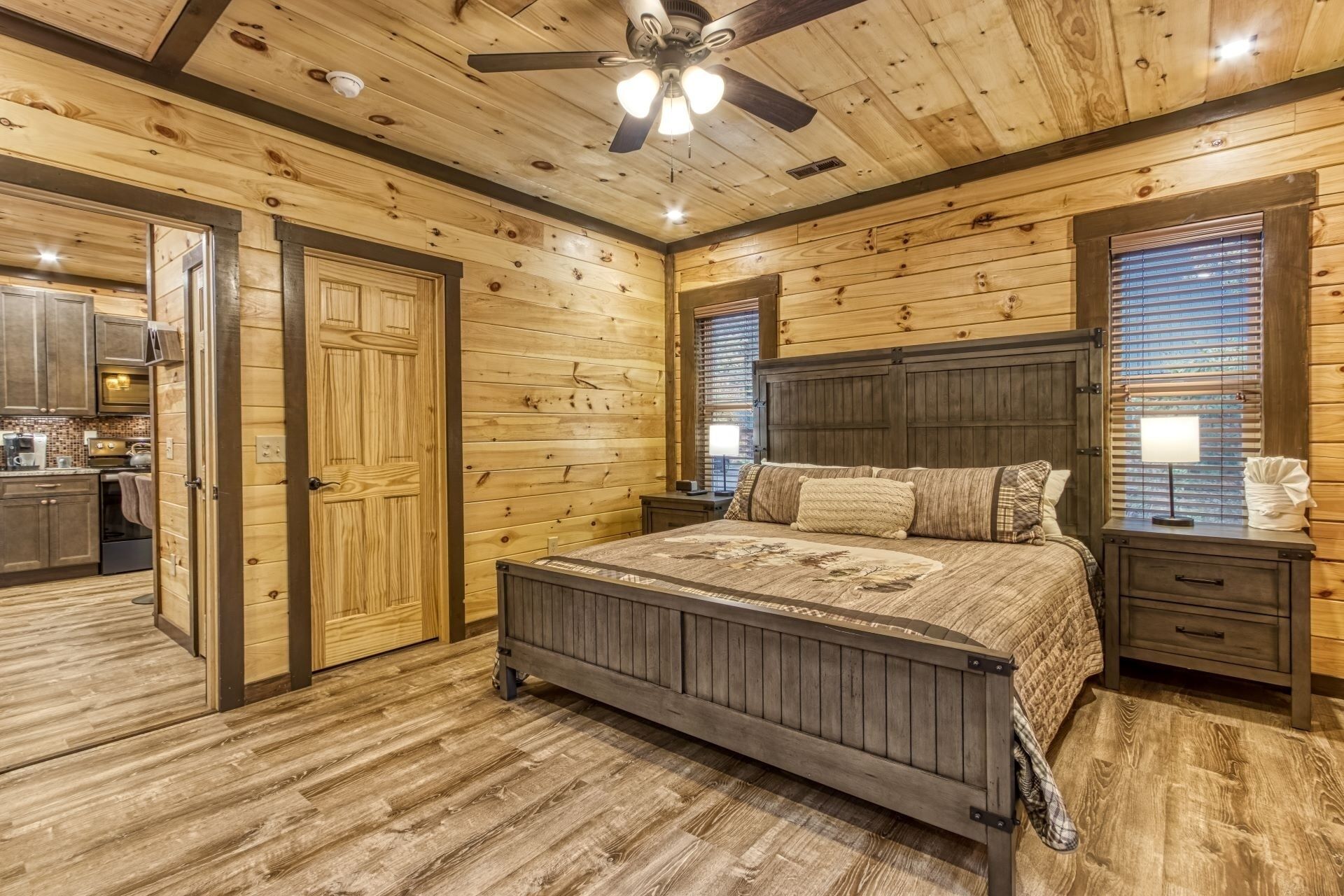 Un dormitorio de estilo cabaña de troncos con una cama de madera, un ventilador de techo y vistas a la cocina a través de una ventana.