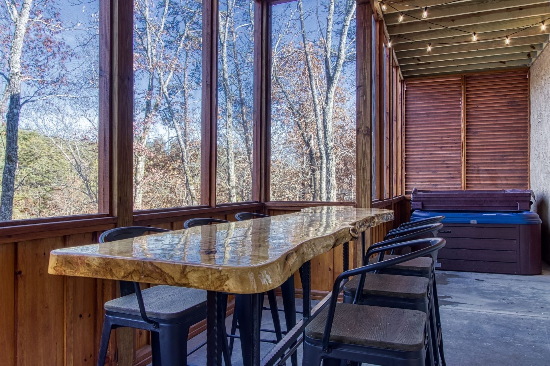 A rustic wooden interior with a large table and chairs, a hot tub, and large windows offering a view of trees.
