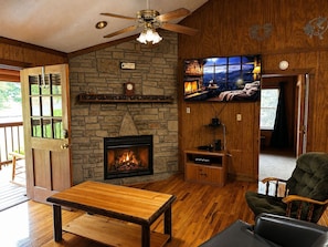 Ein gemütliches Wohnzimmer mit Kamin aus Stein, Holzboden, Deckenventilator und einem an der Wand befestigten Fernseher.