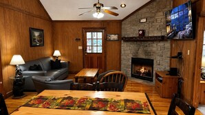 A cozy living room with a fireplace, wooden walls, a sofa, a dining table with a placemat, and a ceiling fan. - Er31 - Grandma And Granpaws Place - Great Location - Close To Parkway In Pf 2 Bedroom Cabin by RedAwning (Pigeon Forge)