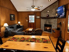 A cozy living room with a fireplace, wooden walls, a sofa, a dining table with a placemat, and a ceiling fan. - Er31 - Grandma And Granpaws Place - Great Location - Close To Parkway In Pf 2 Bedroom Cabin by RedAwning (Pigeon Forge)