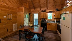 A wooden cabin interior with a dining table, chairs, a kitchen with wooden cabinets, a refrigerator, and a window with blinds. - Er75 - Palmetto Place - Great Location! Close To All The Action! 2 Bedroom Cabin by RedAwning (Pigeon Forge)