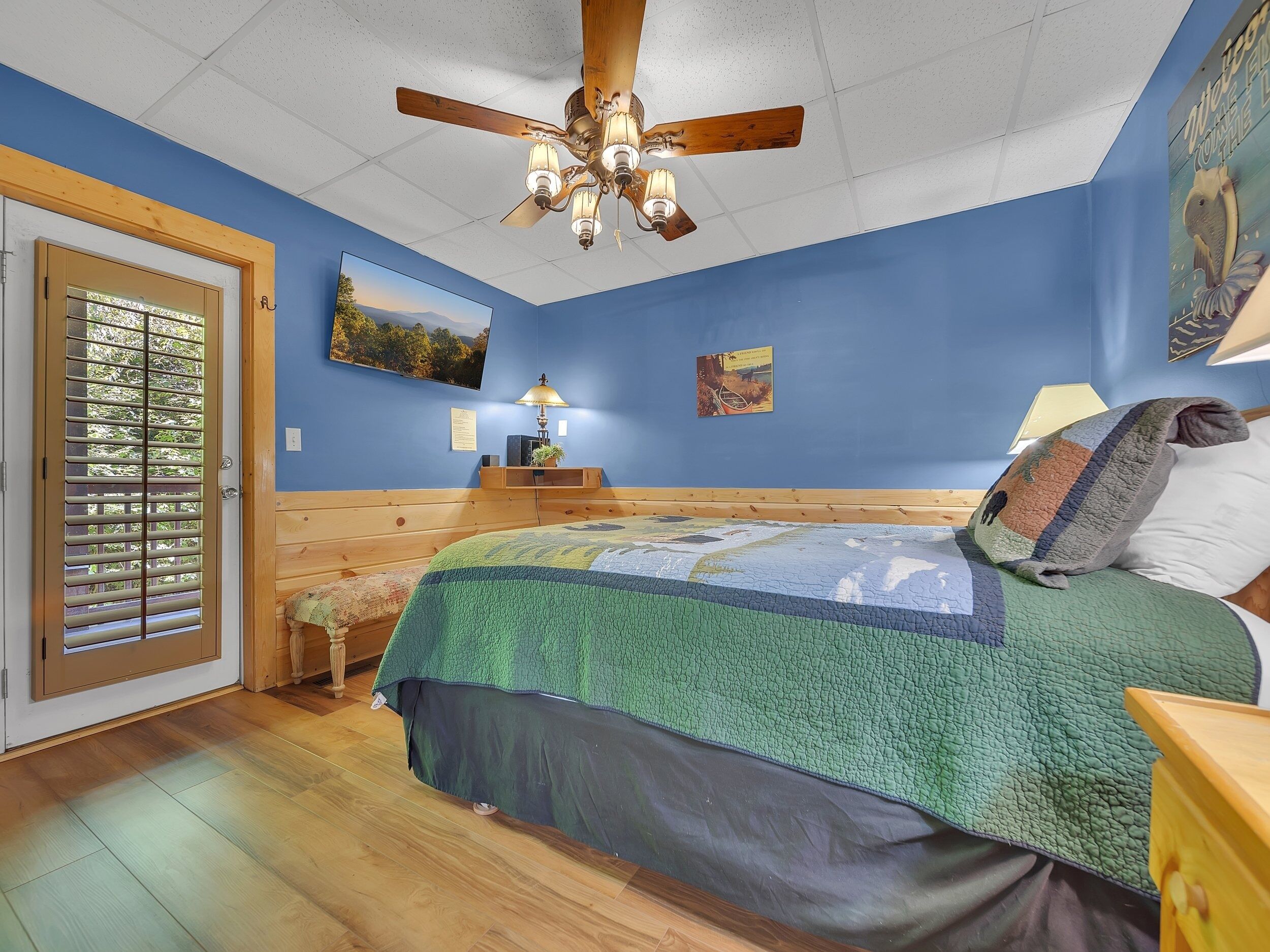 A bedroom with a wooden bed, a ceiling fan, a door with blinds, and a wall-mounted lamp.