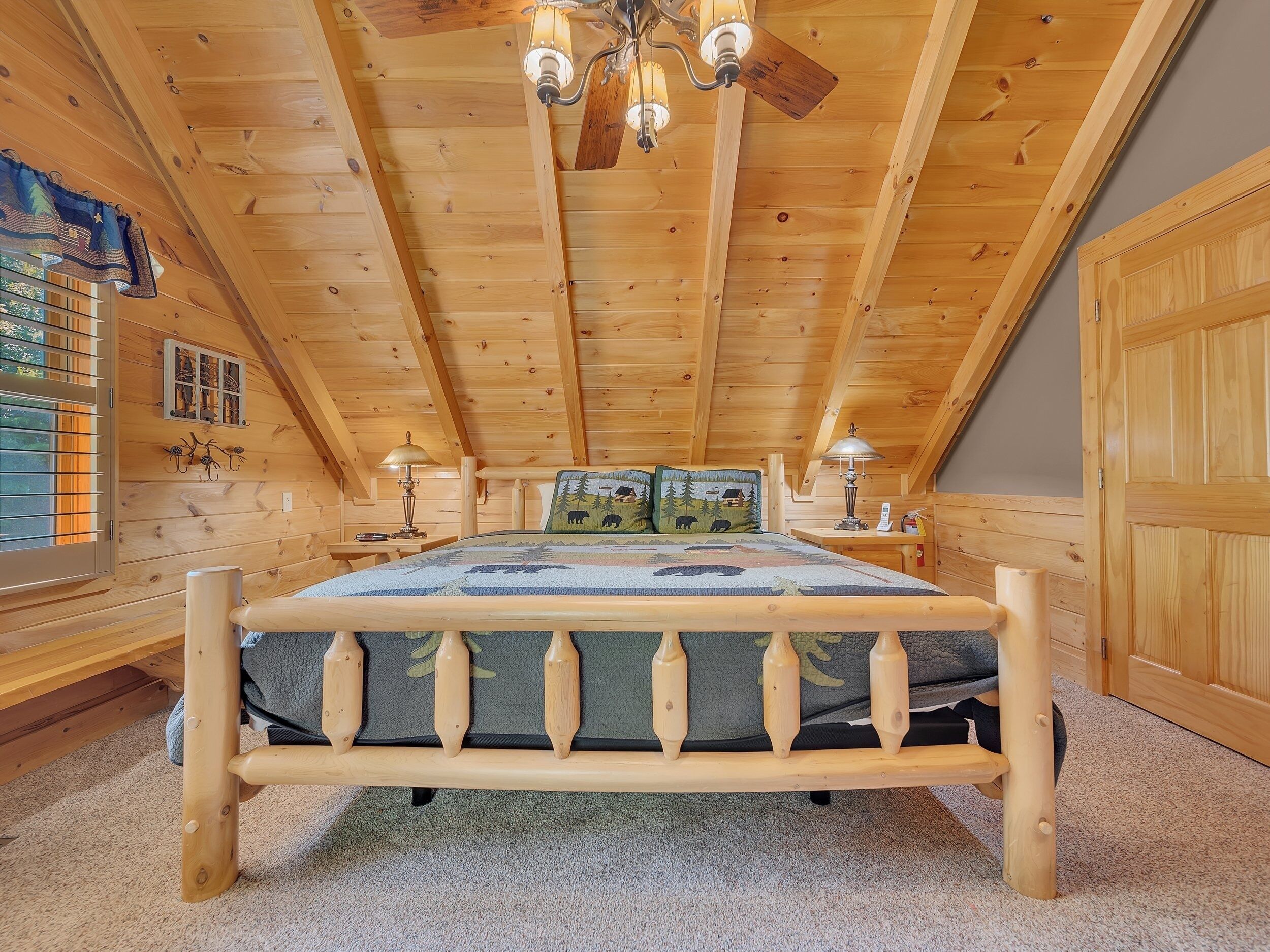 A wooden cabin bedroom with a bed, two bedside tables, a ceiling fan, and a window with blinds.