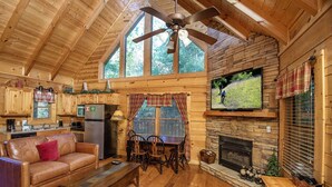 A cozy cabin interior with a wooden ceiling, a stone fireplace, a television, and a dining area. - ER244 - Black Hawk Hideaway Great Location - Close To Town! 2 Bedroom Cabin by RedAwning (Pigeon Forge)
