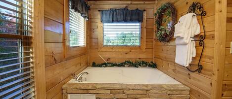 A wooden cabin bathroom with a stone bathtub, wooden walls, and a window with blinds.