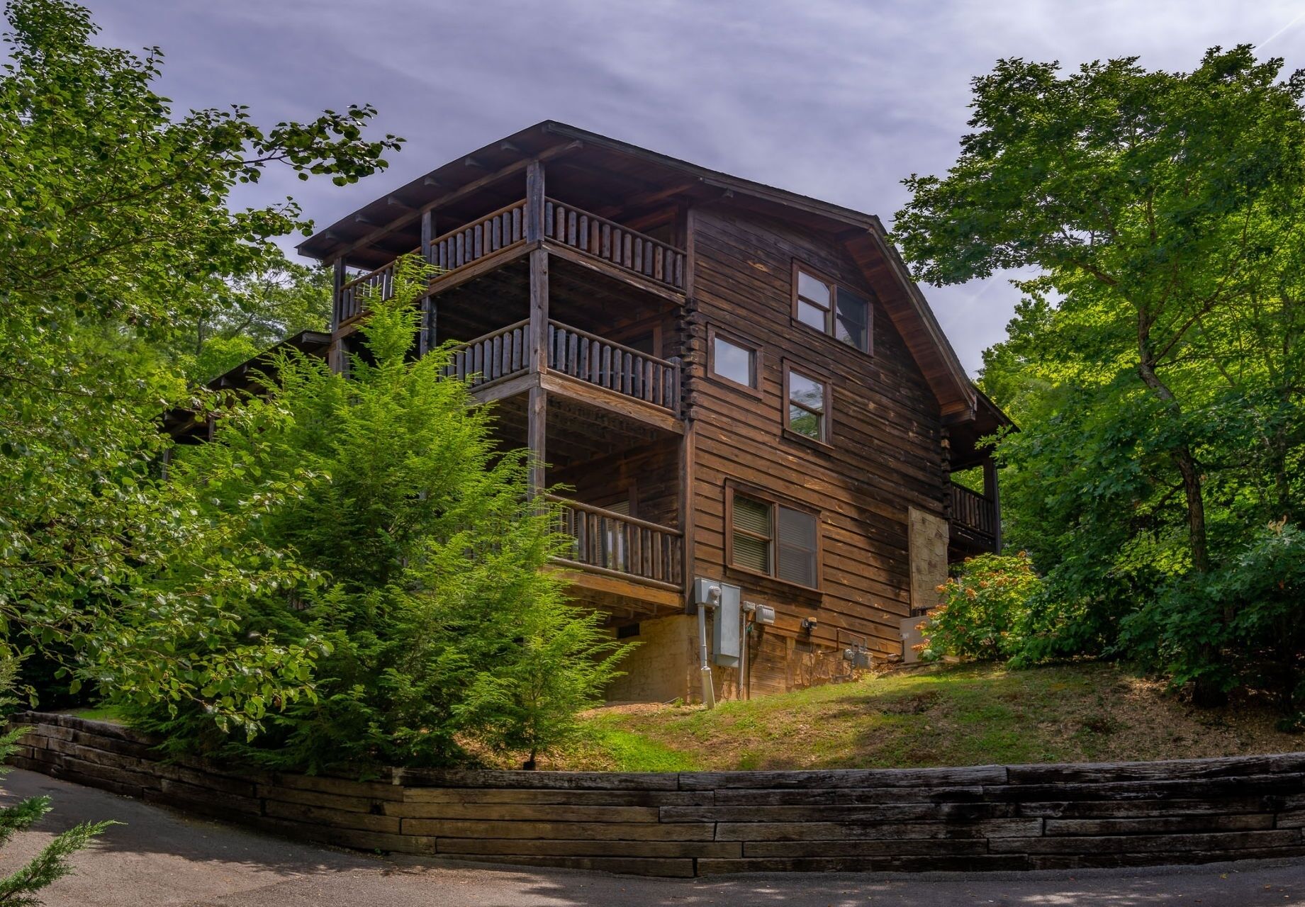 A multi-story log cabin with a balcony on the upper floor, surrounded by dense greenery.