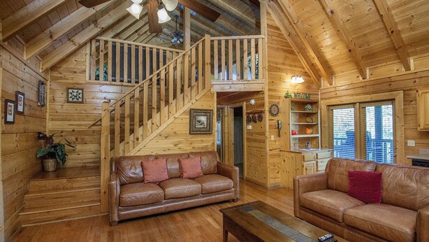 A wooden cabin interior with a staircase leading to a loft, two leather sofas, a wooden coffee table, and a ceiling fan. - ER244 - Black Hawk Hideaway Great Location - Close To Town! 2 Bedroom Cabin by RedAwning (Pigeon Forge)