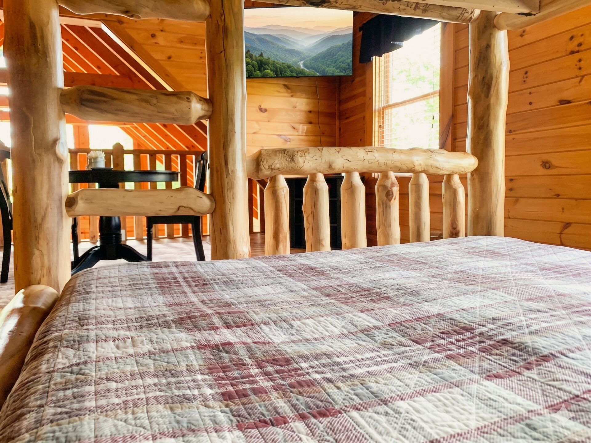 A wooden cabin interior with a bed, a chair, and a view of a forest through a window.