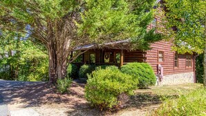 A rustic log cabin with a porch, surrounded by dense greenery and trees. - Er1 - Smoky Mountain Escape Great Location - Close To Town! 3 Bedroom Cabin by RedAwning (Pigeon Forge)