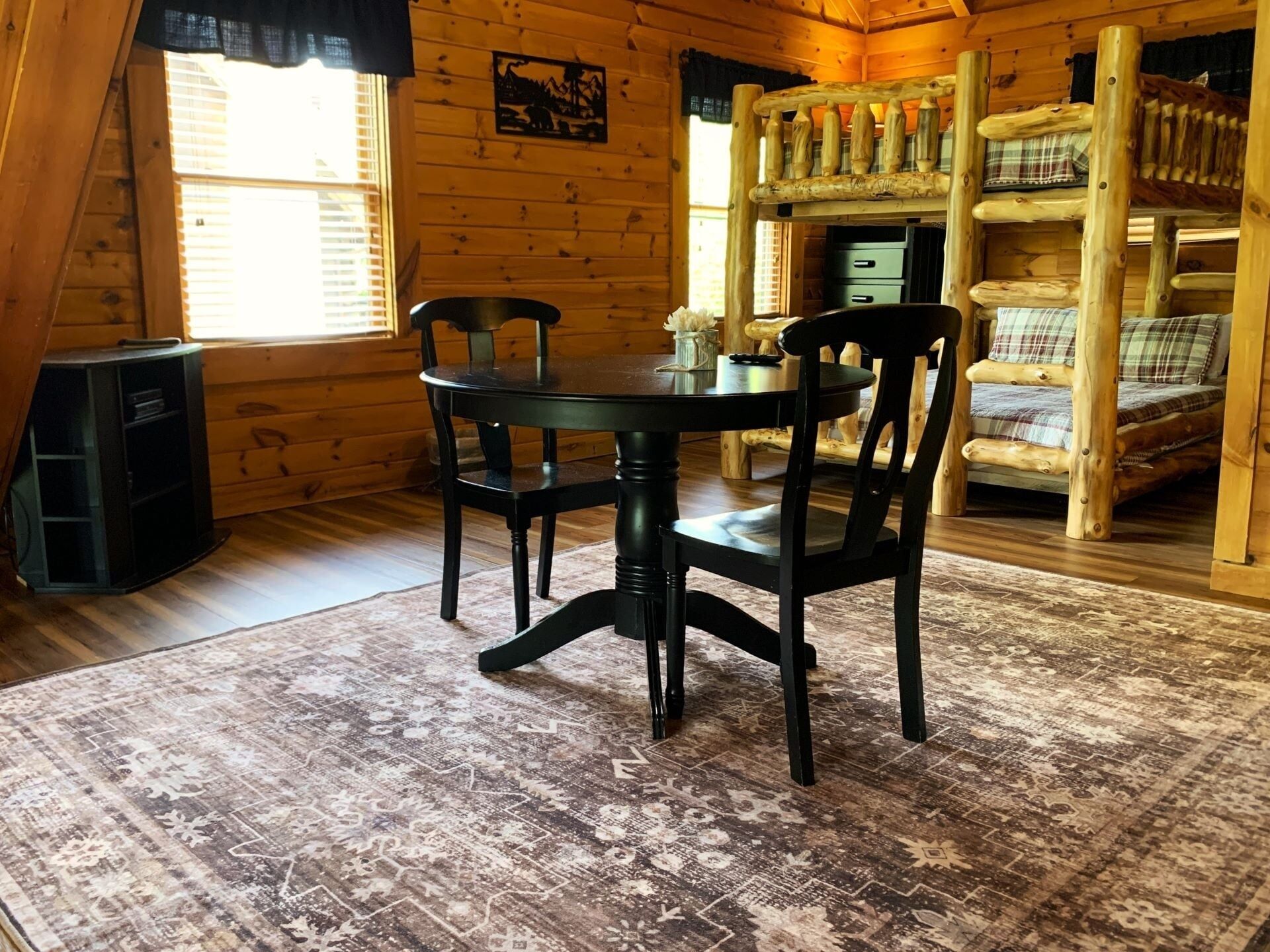 A wooden cabin interior with a dining table, chairs, a bunk bed, and a speaker.