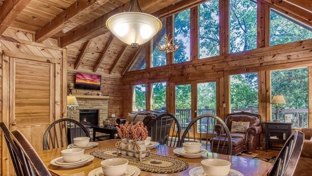 A wooden cabin interior with a dining table set for six, a fireplace, and large windows offering a view of trees. - Er1 - Smoky Mountain Escape Great Location - Close To Town! 3 Bedroom Cabin by RedAwning (Pigeon Forge)