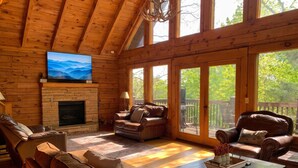 A log cabin interior with a fireplace, leather sofas, a television, and large windows. - Er1 - Smoky Mountain Escape Great Location - Close To Town! 3 Bedroom Cabin by RedAwning (Pigeon Forge)