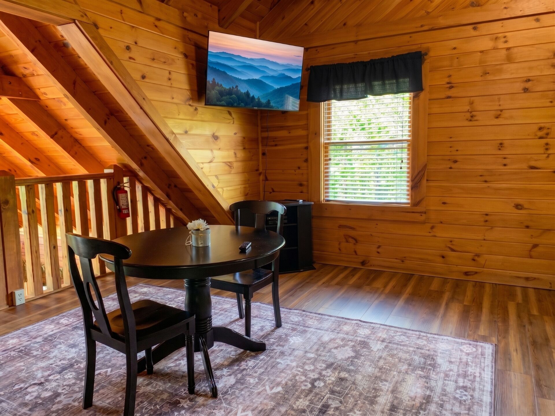 Une cabane en bois comprenant un coin repas avec une table et des chaises, un téléviseur fixé au mur et une fenêtre avec des stores.