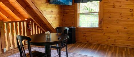 Une cabane en bois comprenant un coin repas avec une table et des chaises, un téléviseur fixé au mur et une fenêtre avec des stores.