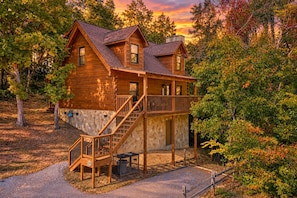 A two-story log cabin with a stone foundation, a wooden deck, and a staircase leading to the upper level. - Er27- Bear Naked - Great Location- Close To Town 2 Bedroom Cabin by RedAwning (Pigeon Forge)