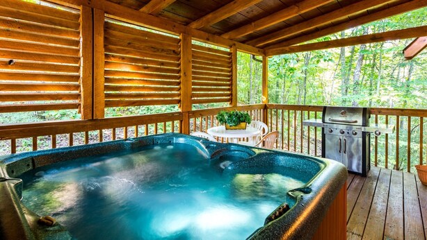 A wooden deck with a hot tub, a grill, and a table with chairs. - Er27- Bear Naked - Great Location- Close To Town 2 Bedroom Cabin by RedAwning (Pigeon Forge)