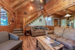 A log cabin interior with a staircase, two sofas, a coffee table, and a kitchen area.