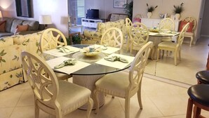 A dining area with a round glass table, white chairs, and a floral-patterned sofa. - Sea Place 13137 2 Bedroom Condo by RedAwning (St. Augustine)