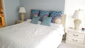 A bedroom with a bed, two bedside tables, a wall decoration, and a window with blinds. - Sea Place 13137 2 Bedroom Condo by RedAwning (St. Augustine)