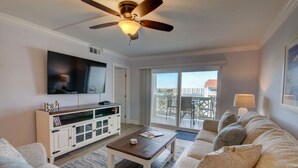 A living room with a ceiling fan, a flat-screen TV, a white entertainment unit, a sofa, a coffee table, and a balcony with a view of buildings. - 461 El Matador 2 Bedroom Condo by RedAwning (Fort Walton Beach)