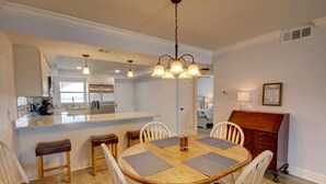 A dining area with a wooden table and white chairs, a chandelier, and a kitchen in the background. - 461 El Matador 2 Bedroom Condo by RedAwning (Fort Walton Beach)