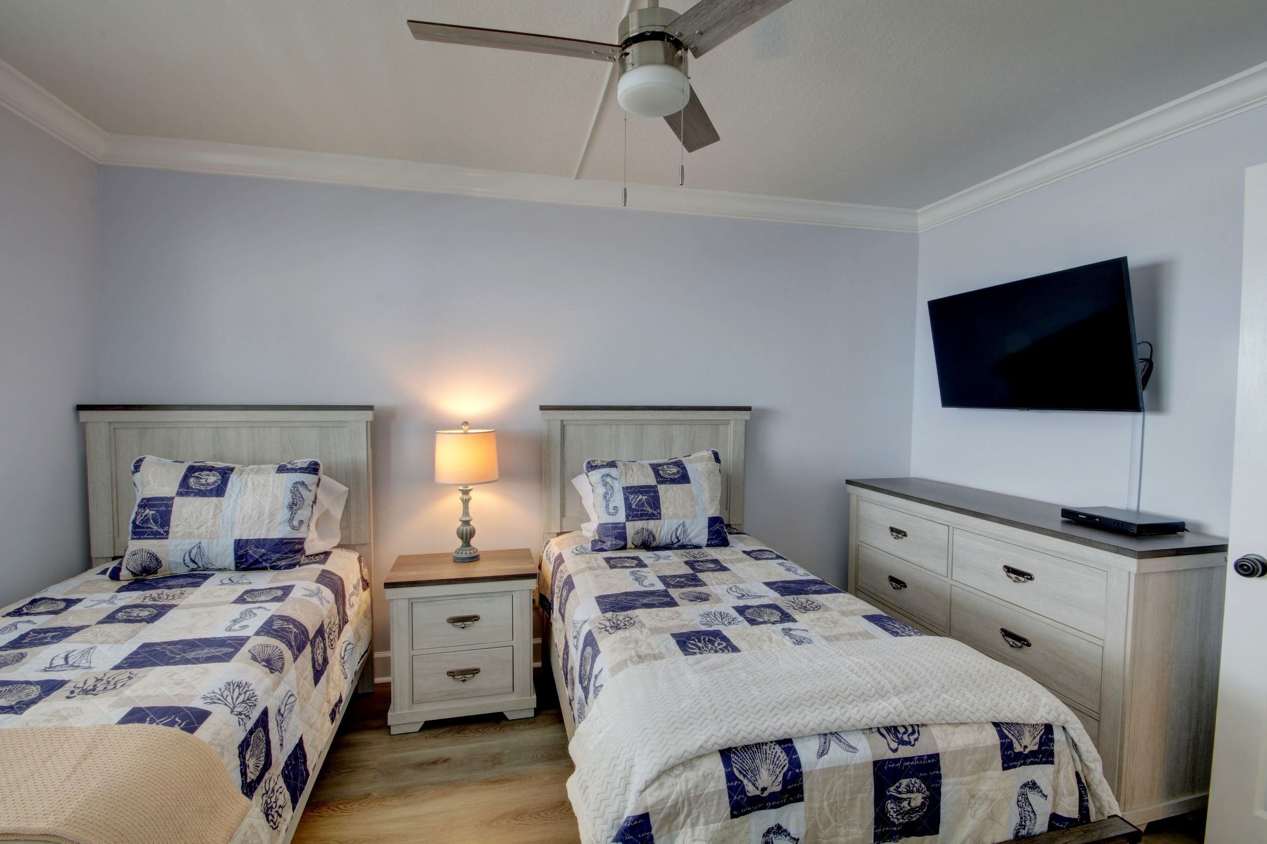 A bedroom with two beds, a ceiling fan, a TV mounted on the wall, and a bedside table with a lamp.