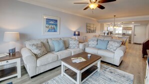 A living room with a sectional sofa, a coffee table, a ceiling fan, and a kitchen in the background. - 461 El Matador 2 Bedroom Condo by RedAwning (Fort Walton Beach)