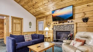 A cozy living room with a stone fireplace, a leather armchair, a blue sofa, and a television mounted on the wall. - Er202 - Now And Forever Great Location! - Close To Town! 4 Bedroom Cabin by RedAwning (Pigeon Forge)