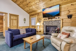 A cozy living room with a stone fireplace, a leather armchair, a blue sofa, and a television mounted on the wall.