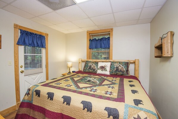 Un dormitorio con cama, dos ventanas con cortinas azules, cabecera de madera, colcha con motivos de osos y una mesita de noche con lámpara.