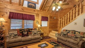 A cozy cabin interior with two sofas adorned with animal-themed cushions, a wooden coffee table, and a staircase leading to an upper level.