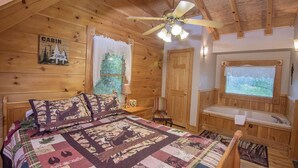A cabin interior with a bed, a window with a view, a ceiling fan, and a wooden door.
