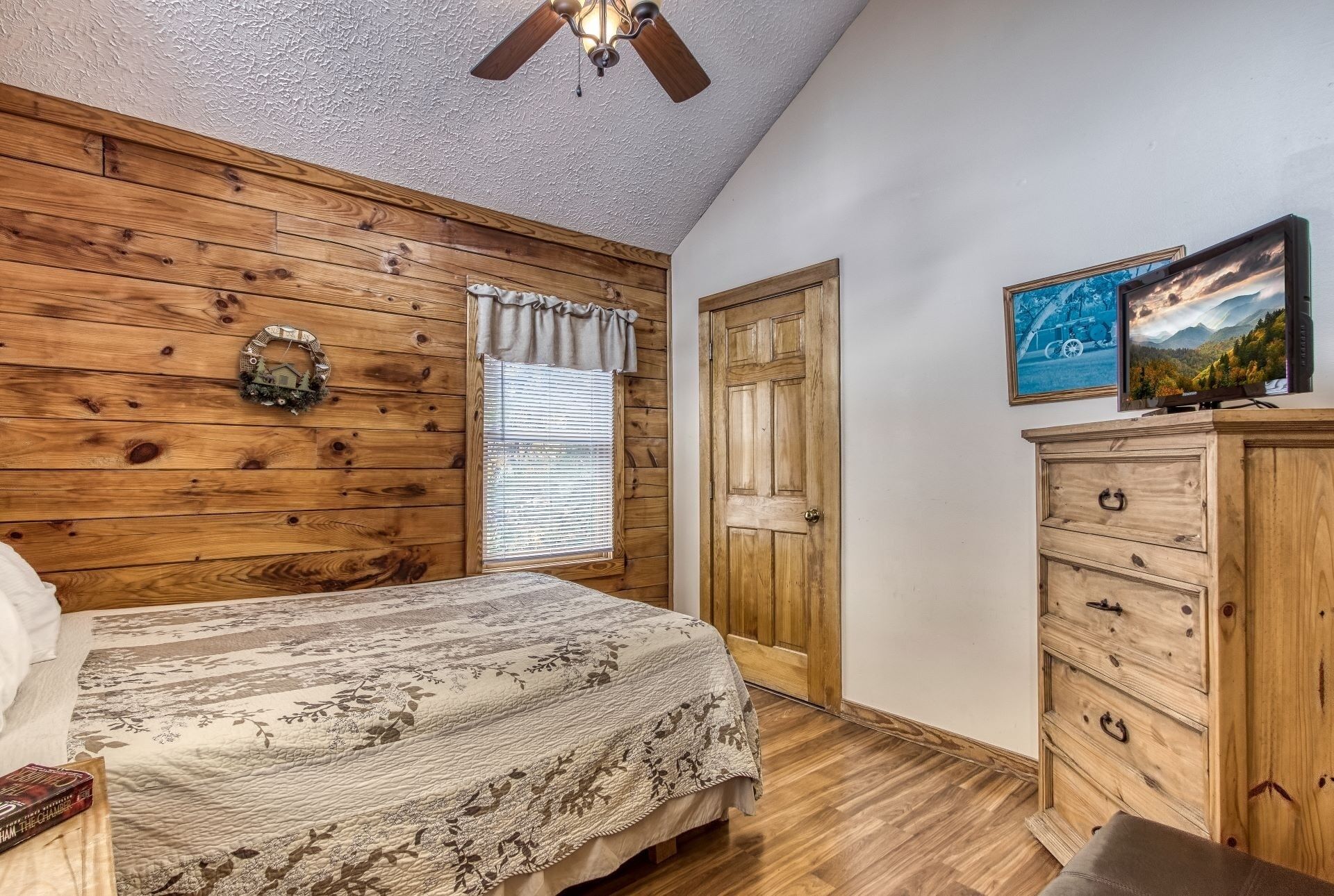 A bedroom with wooden walls, a bed with a floral bedspread, a dresser, and a ceiling fan.