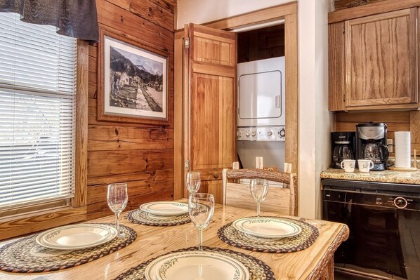 A wooden interior with a dining table set for four, a kitchen with a stove and coffee maker, and a framed picture on the wall. - Er13 - Moose Manor Great Location - Close To Town! 3 Bedroom Cabin by RedAwning (Pigeon Forge)