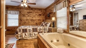 A log cabin bedroom with a large bed, a wooden dresser, a vanity with a mirror, a ceiling fan, and a window with blinds. - Er13 - Moose Manor Great Location - Close To Town! 3 Bedroom Cabin by RedAwning (Pigeon Forge)