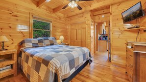 A wooden cabin bedroom with a bed, a ceiling fan, a TV, and a view of the outdoors through a window. - Tranquility - Quiet And Comfortable Cabin! Close To The Tanger Outlet Shopping 2 Bedroom Cabin by RedAwning (Sevierville)