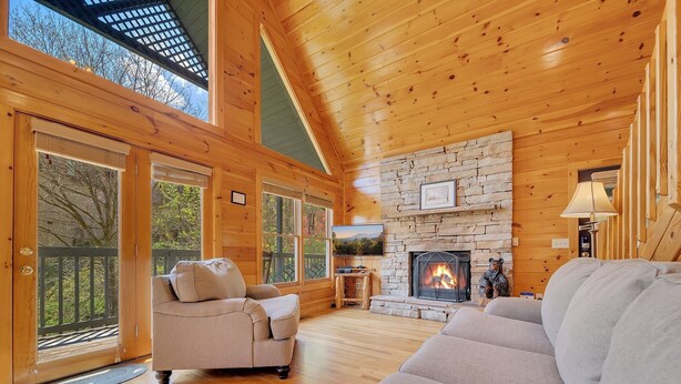 A cozy cabin interior with a stone fireplace, wooden floors, and a large window overlooking a deck. - Tranquility - Quiet And Comfortable Cabin! Close To The Tanger Outlet Shopping 2 Bedroom Cabin by RedAwning (Sevierville)