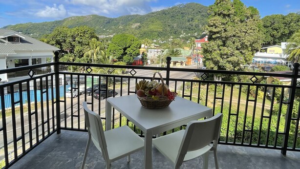 Un balcón con una mesa y sillas, una cesta de mimbre con frutas, y vistas a una montaña y una piscina.