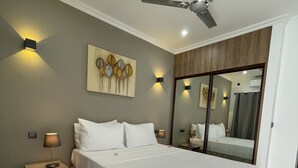 Un dormitorio con una cama grande, un ventilador de techo, una mesita de noche con lámpara, una obra de arte en la pared y un espejo.