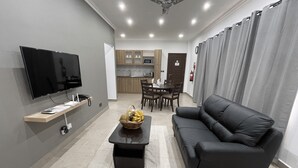 Una sala de estar moderna con un sofá negro, un televisor de pantalla plana, una mesa de centro de madera y un área de comedor al fondo.